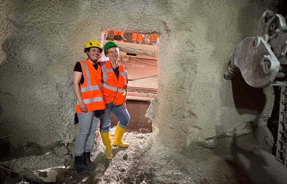 Zwei Frauen in Sicherheitskleidung stehen in einem Tunnel vor einer durchbrochenen Wand in einem Tunnel.